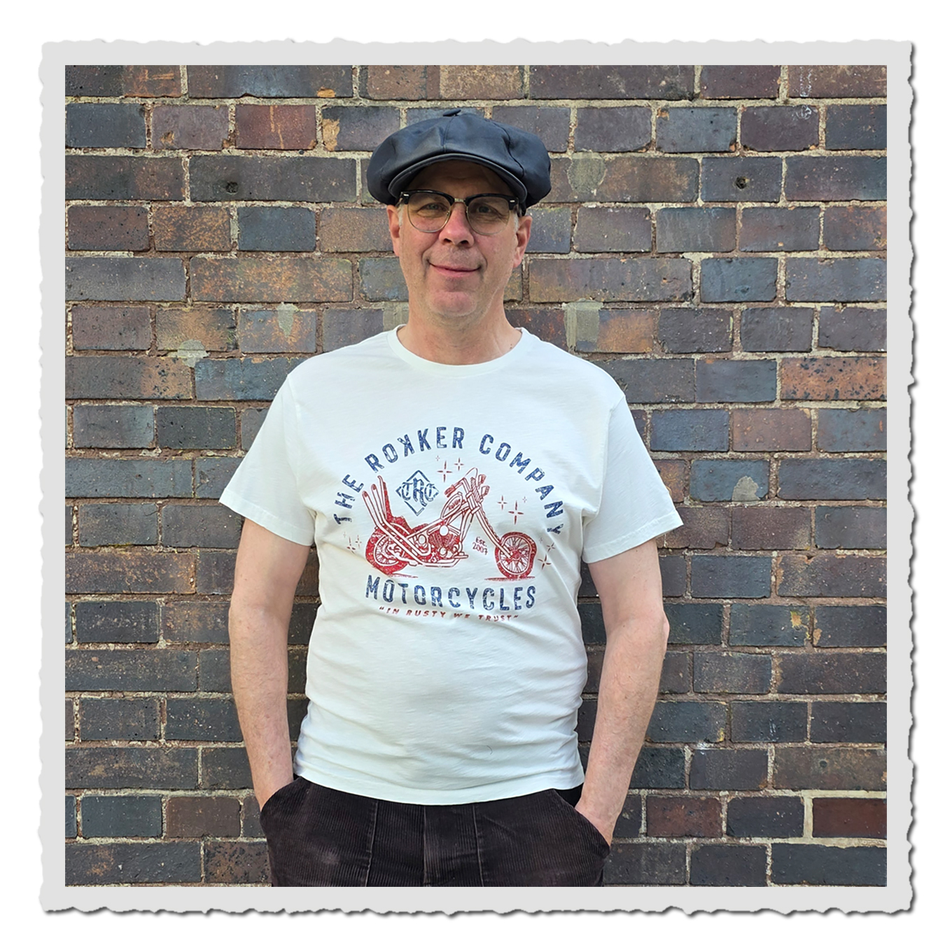 Frontansicht eines Mannes mit heller Shirtgrafik, dunkler Hose und Kappe vor rustikaler Ziegelwand.